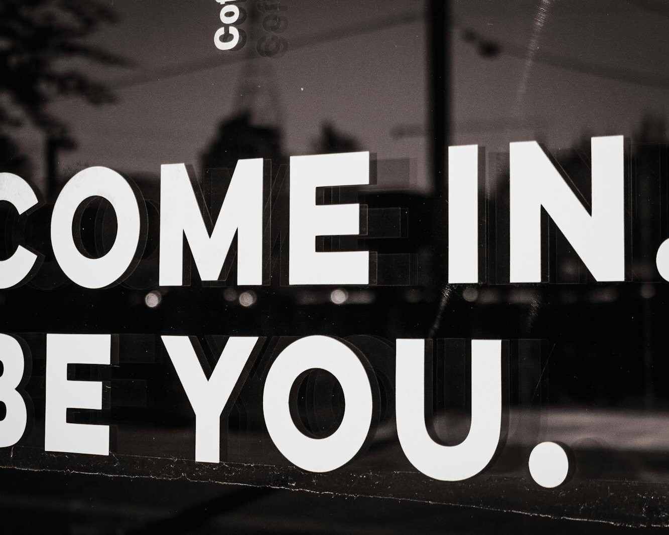 Black and white close-up of a motivational sign on a cafe window inviting customers to be themselves.