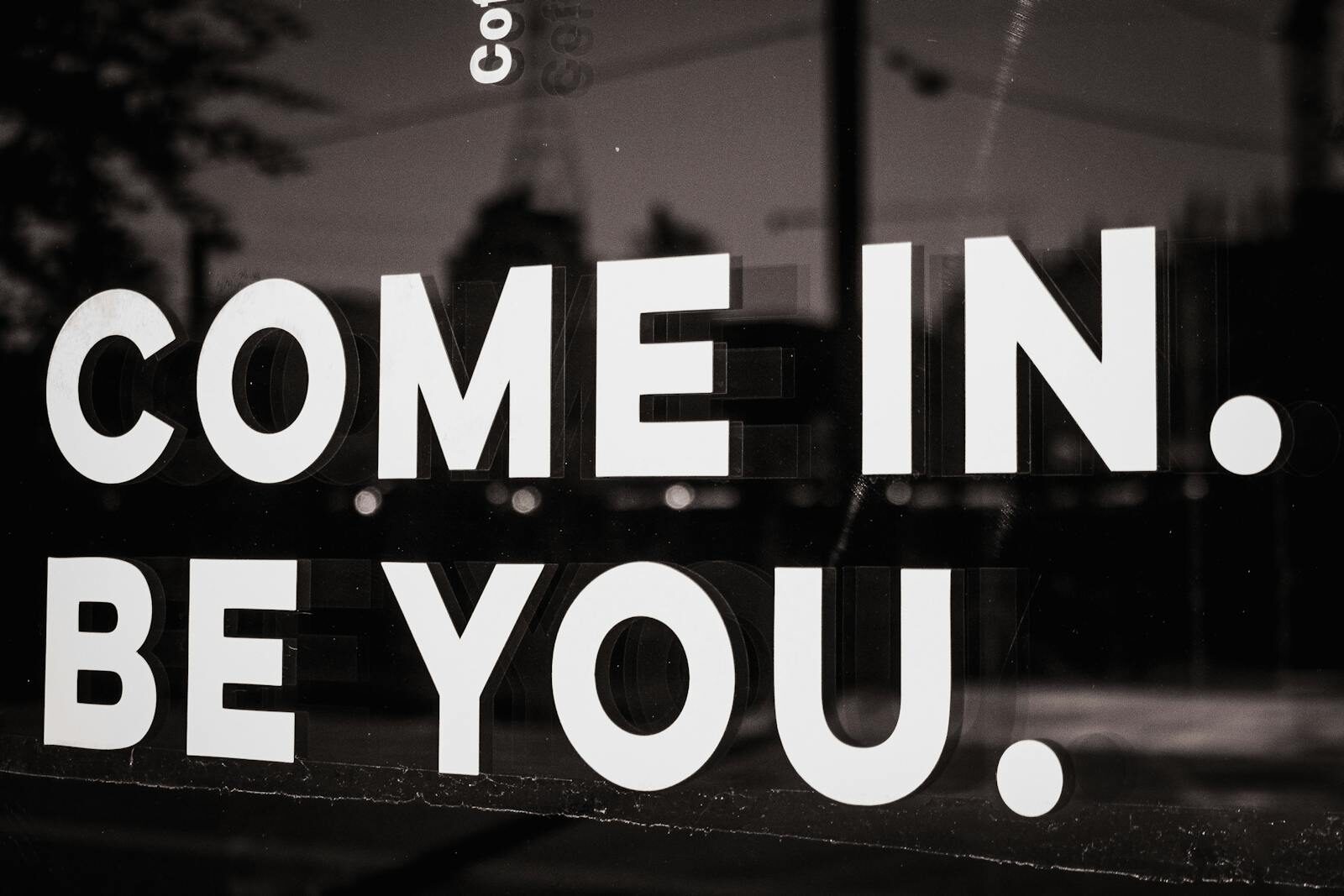 Black and white close-up of a motivational sign on a cafe window inviting customers to be themselves.